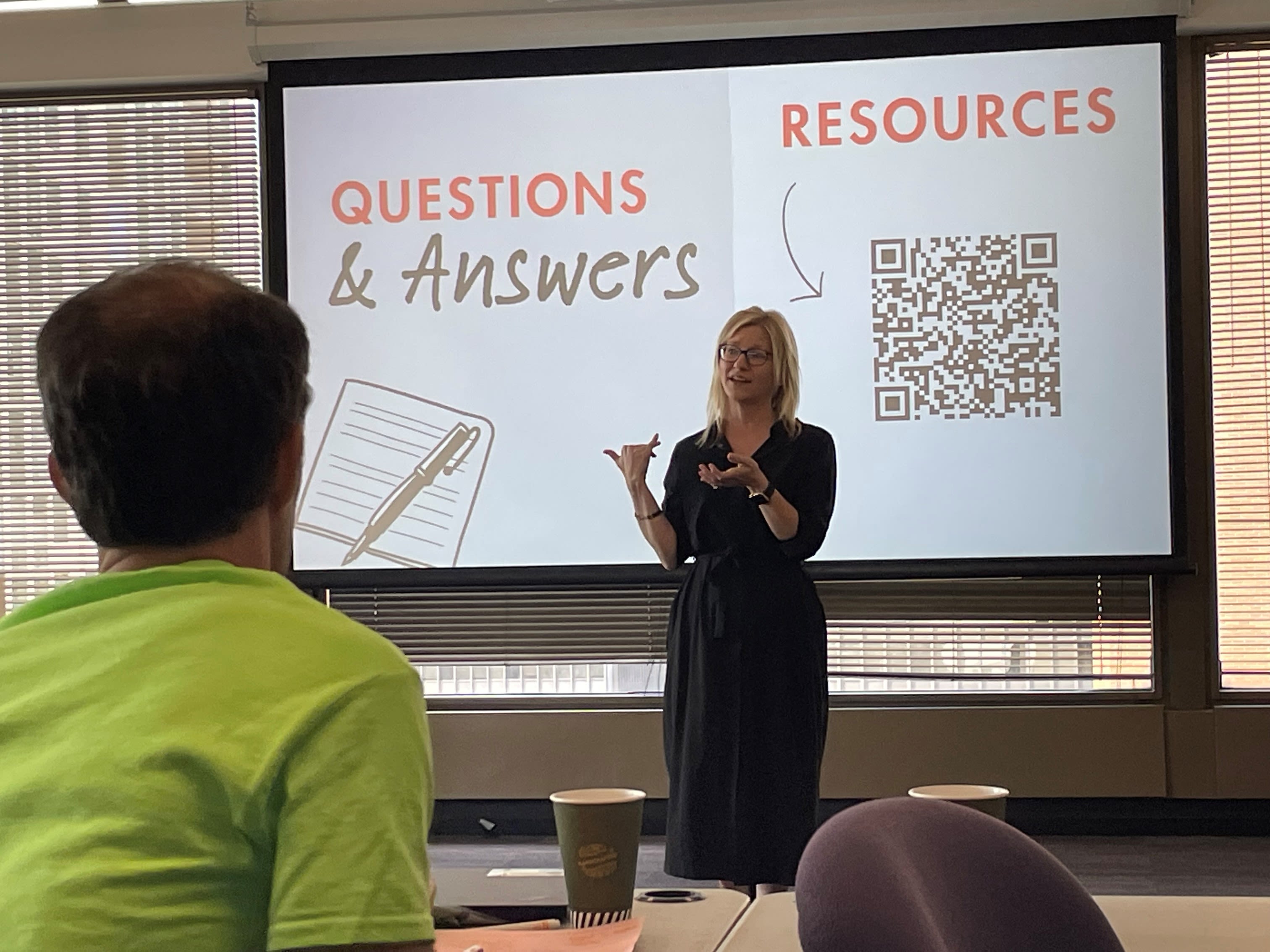 A person in a black dress gives a presentation to others standing in front of a white screen