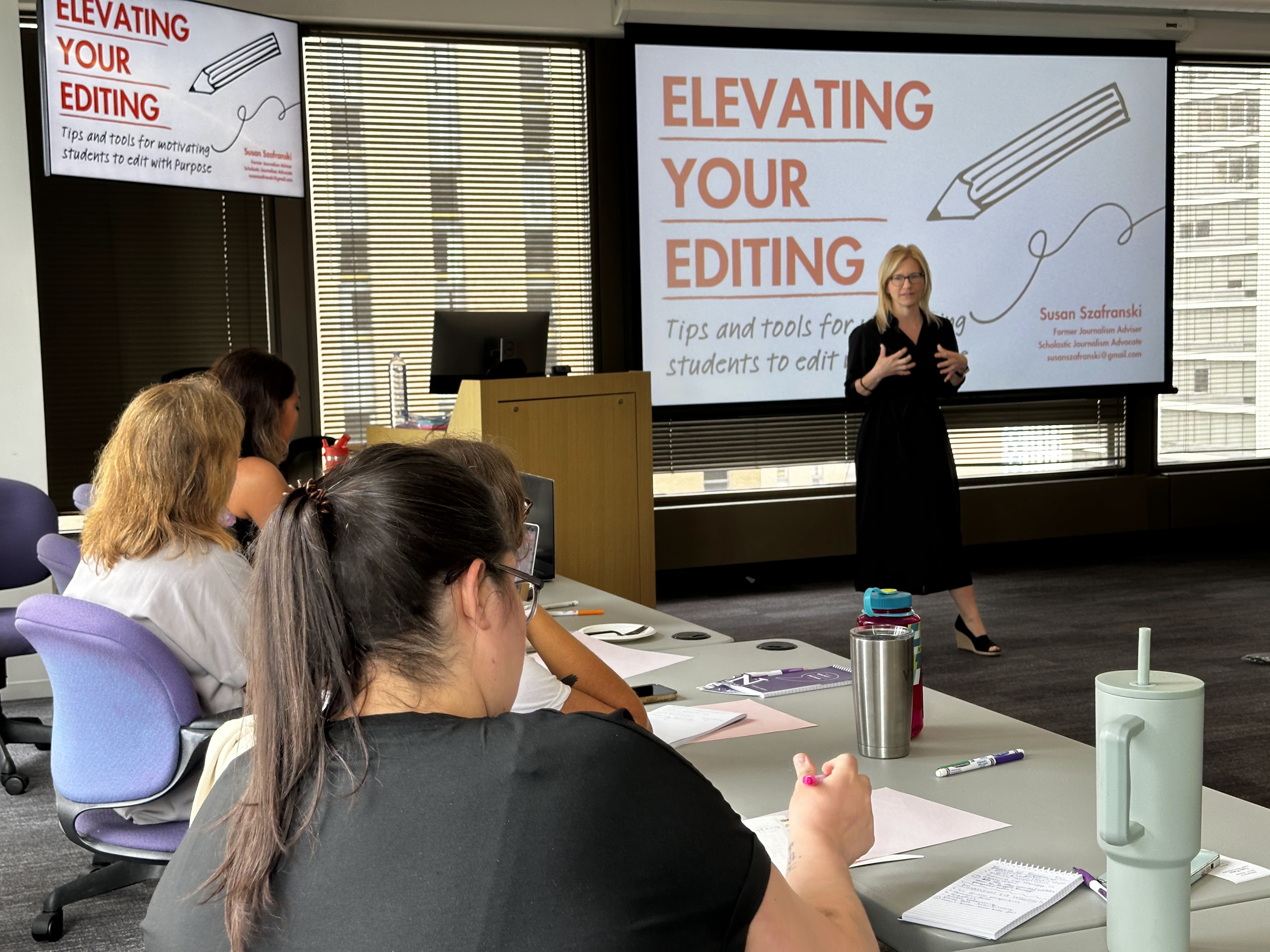 A person in a black dress gives a presentation to others standing in front of a white screen