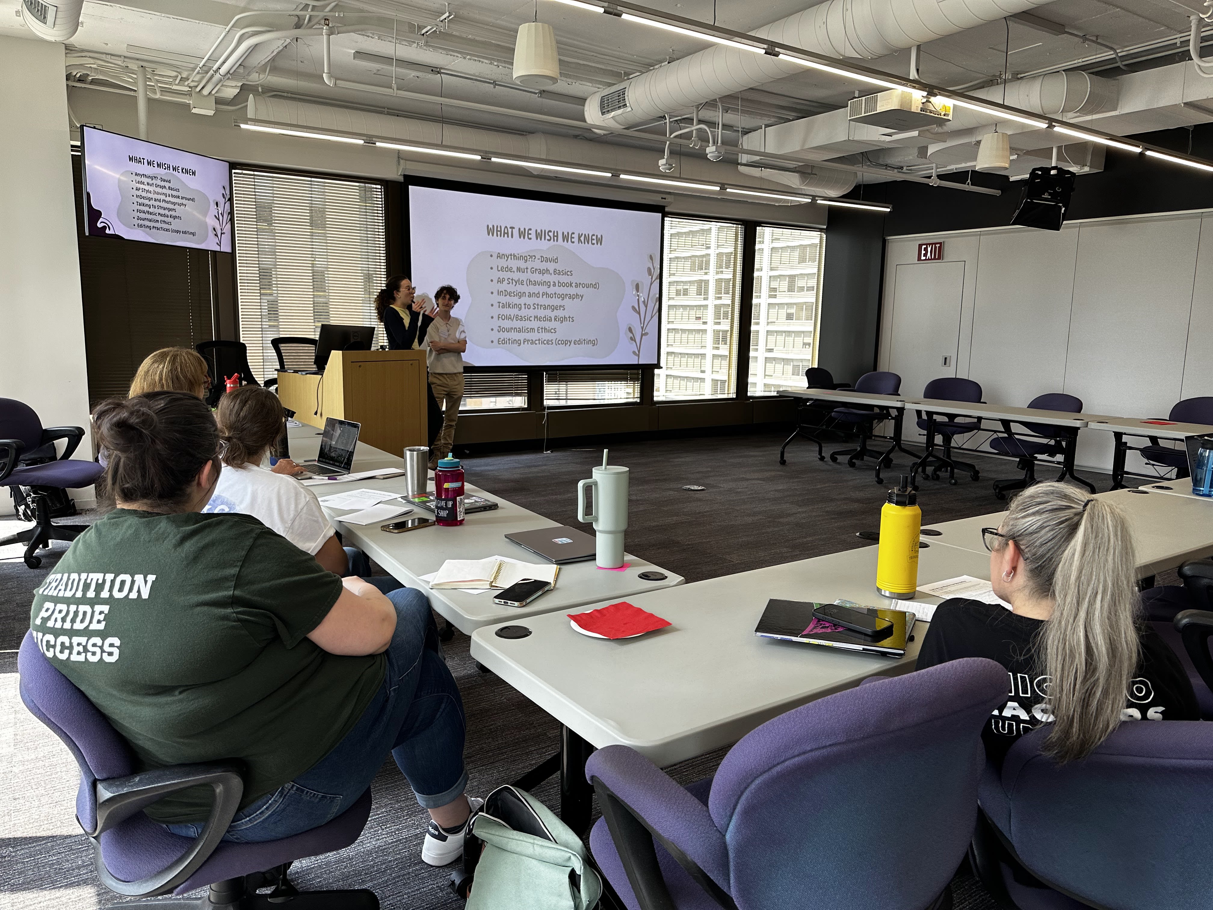 Onlookers sit at a desk and listen to a person giving a presentation