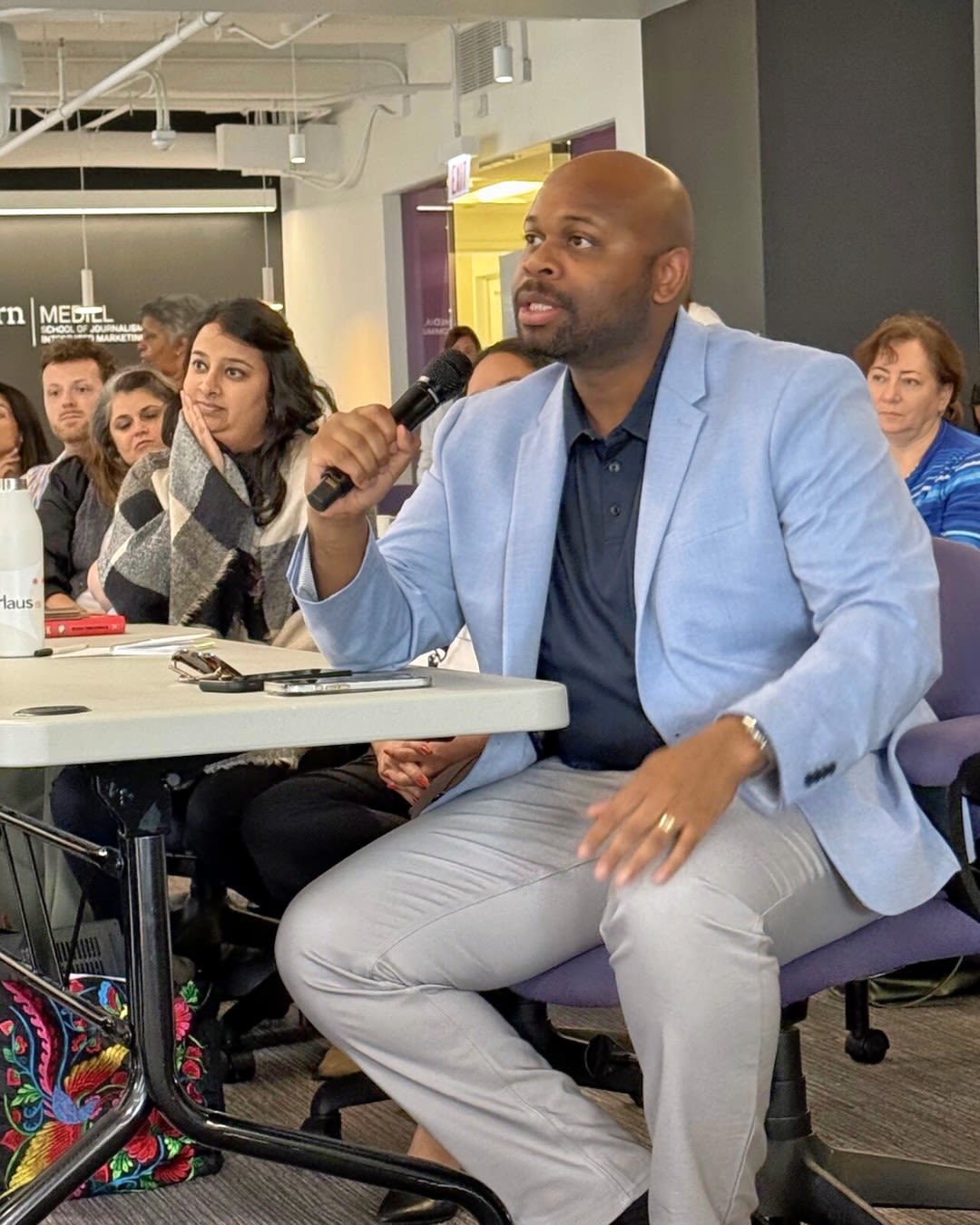 A person in a light blue suit coat and light grey pants holds a microphone and speaks to a room of people
