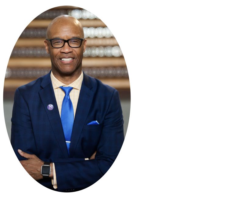 A person, Medill Dean Charles Whitaker, wears glasses and a dark blue suit with a blue tie and white dress shirt as he stands smiling with arms casually crossed