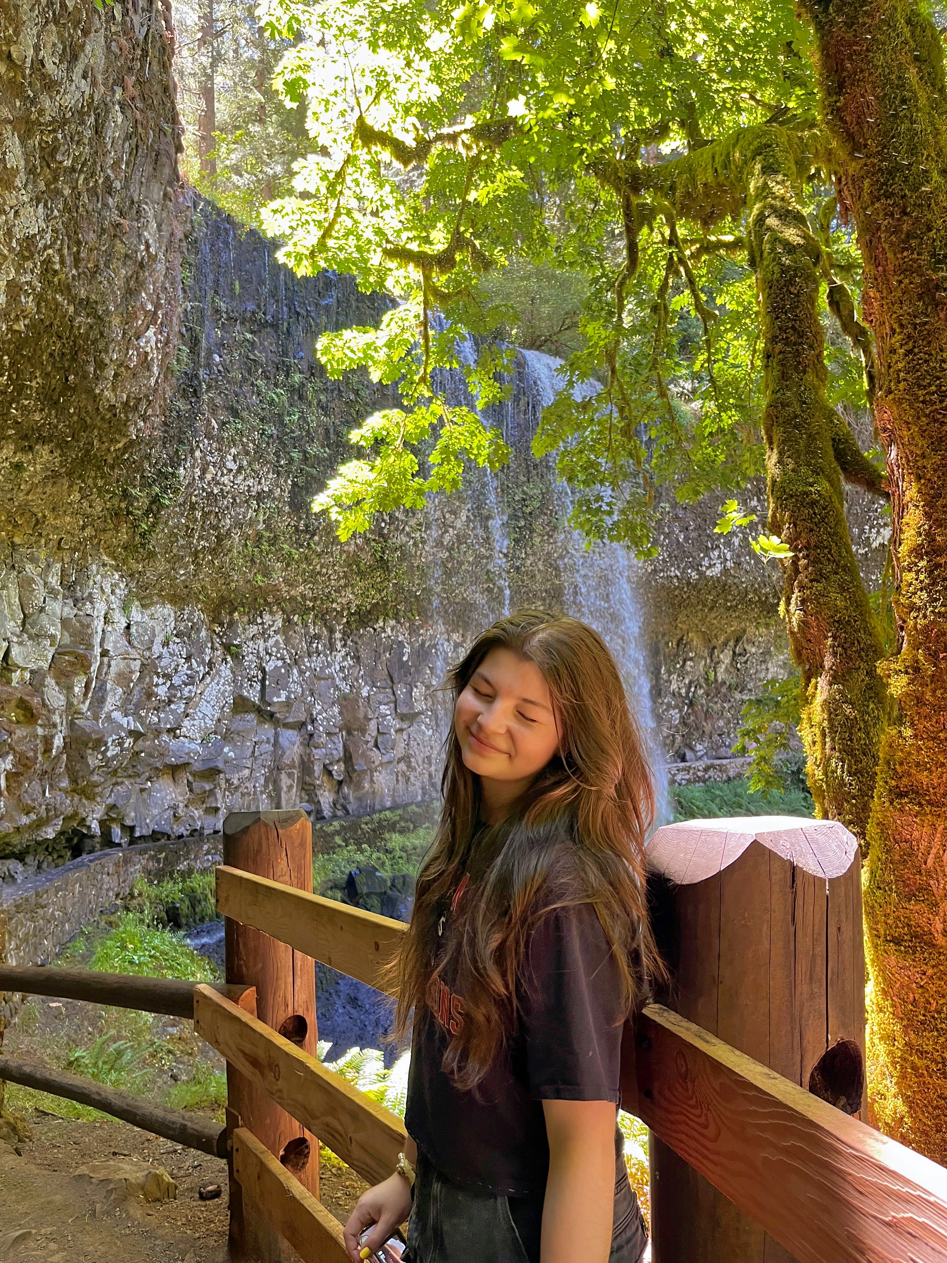 A young person with long dark hair stands with her eyes closed while smiling in front of a steep rock wall in sunlight