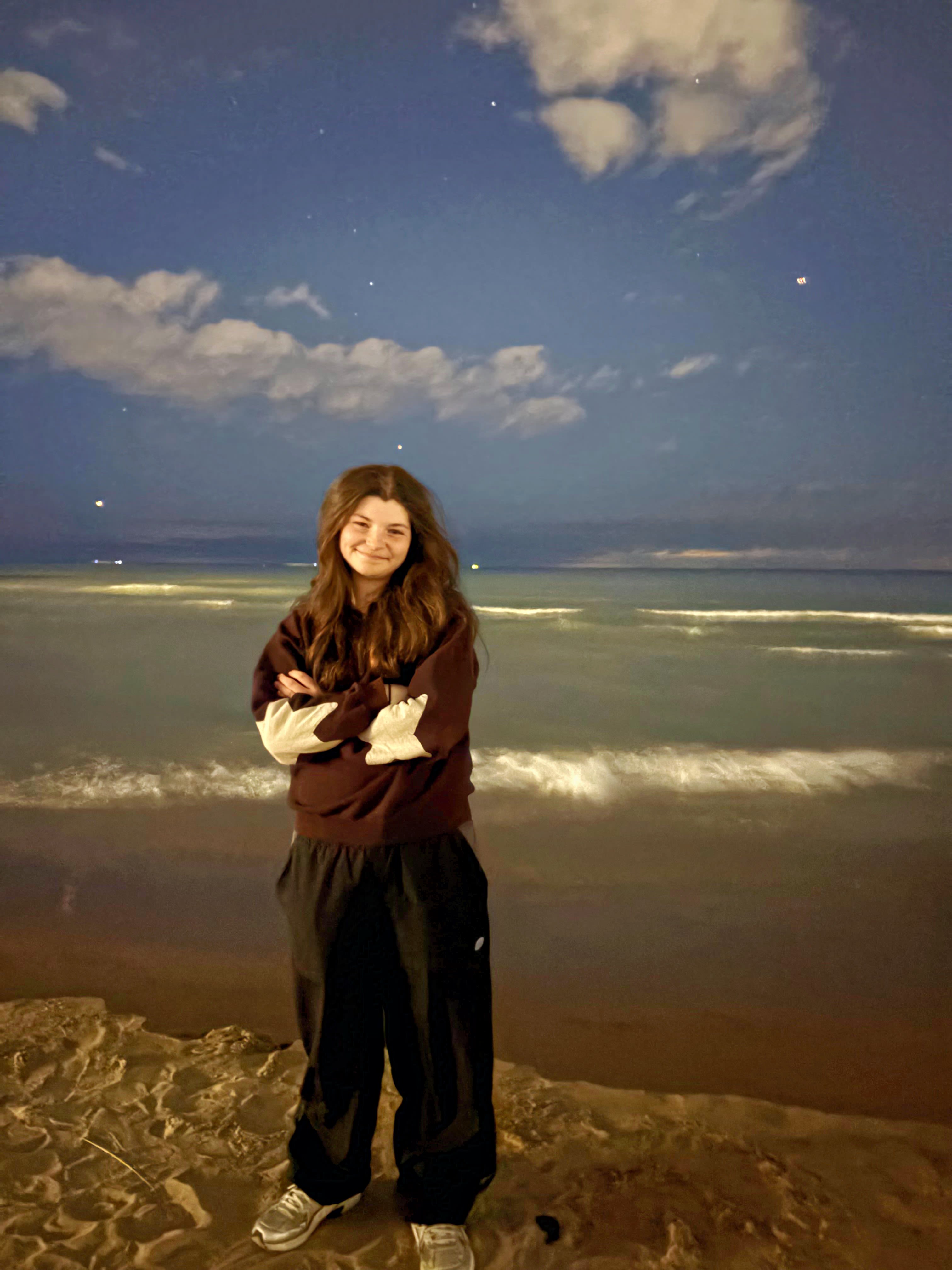 A smiling young person with long dark hair stands with her arms crossed, wearing a casual outfit, along a shore