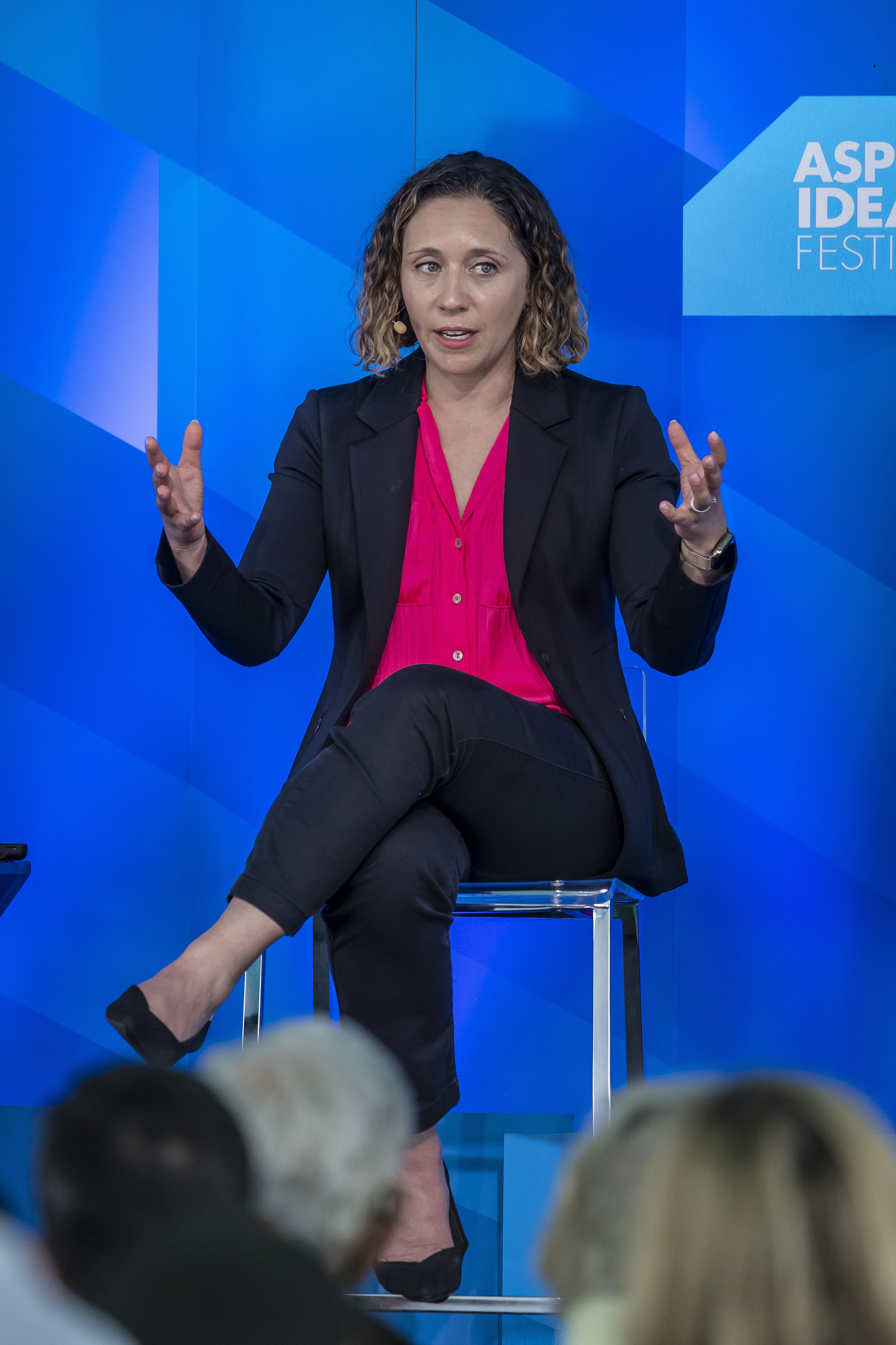 A person, Julie Pace, wearing a pink blouse and dark suit speaks to an audience while sitting in a chair