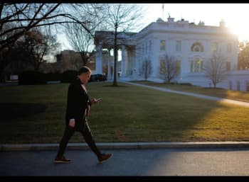 A person, Peter Alexander, walks next to the White House while also looking down at a phone