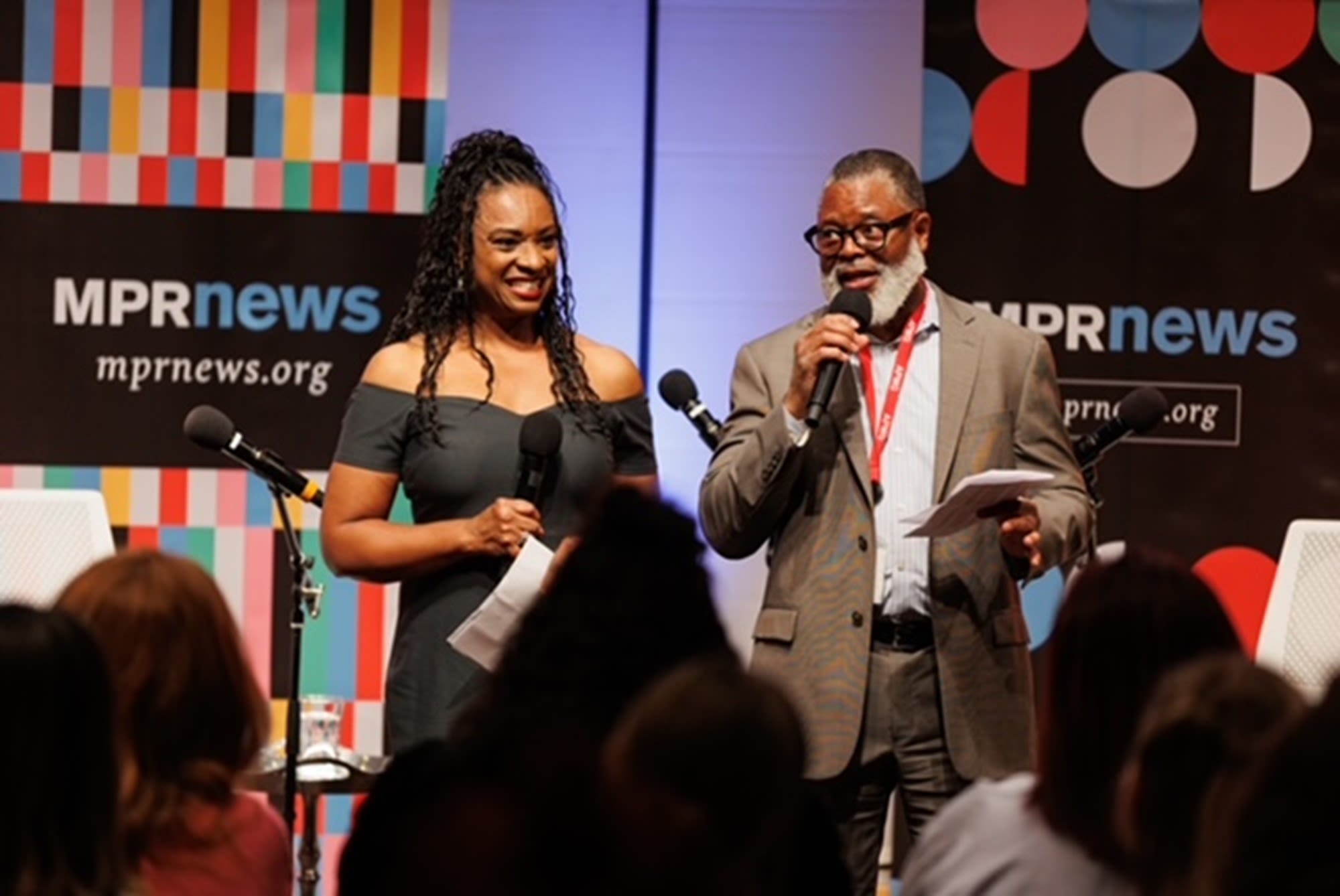 Two people stand onstage and address an audience in front of MPR News banners
